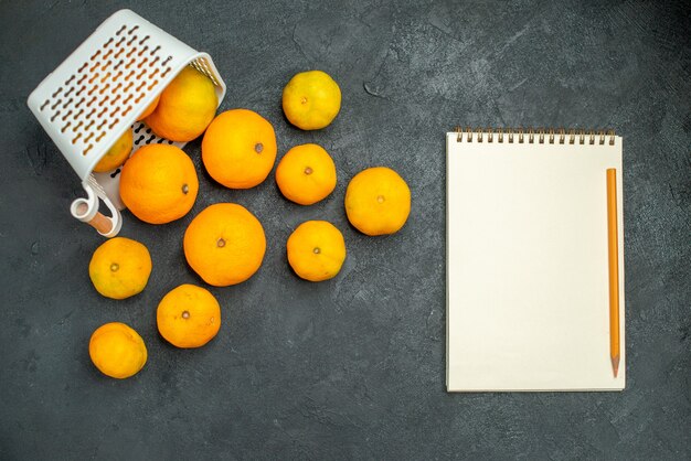 Vista superior de mandarinas y naranjas esparcidas desde el lápiz de cuaderno de cesta de plástico sobre fondo oscuro