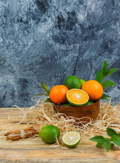 Vista superior de mandarinas en tazón de madera con canela, limas y hojas sobre tablero de madera y superficie de mármol azul