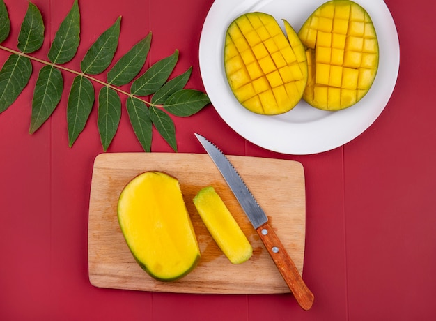 Vista superior de mango picado con cuchillo sobre tablero de cocina de madera con mango en rodajas en un plato blanco con hoja en rojo
