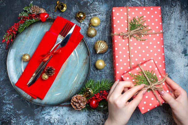Vista superior de la mano abriendo una caja de regalo y cubiertos con cinta roja en una servilleta decorativa en un plato azul y accesorios navideños y calcetín navideño sobre fondo oscuro