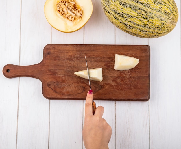 Foto gratuita vista superior de la mano femenina cortando melón en rodajas con un cuchillo en el tablero de cocina de madera en madera blanca