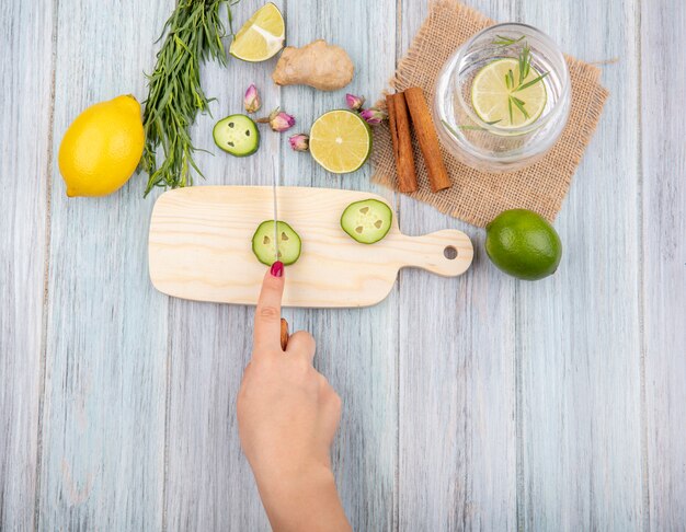 Vista superior de la mano femenina cortando rodajas de pepino en una tabla de cocina de madera con palitos de canela con un vaso de agua en un saco de tela en madera gris