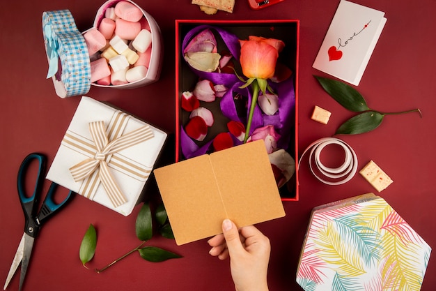 Vista superior de la mano femenina con una pequeña postal abierta sobre la caja de regalo con flor color de rosa color coral con pétalos dispersos y una caja llena de malvaviscos en la mesa roja