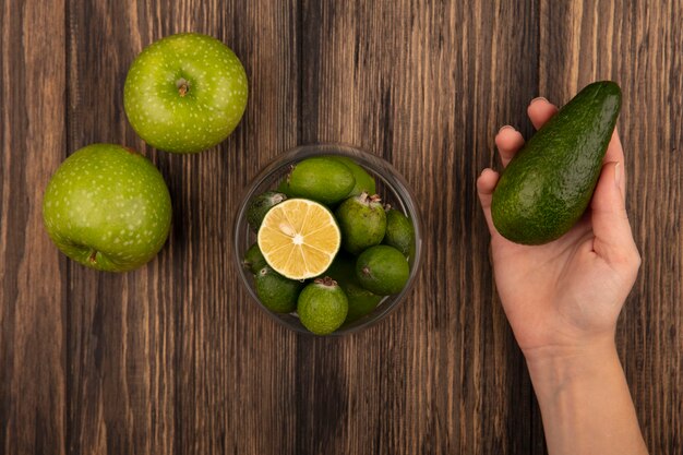 Vista superior de la mano femenina sosteniendo un aguacate fresco con feijoas en un recipiente de vidrio con manzanas verdes aisladas en una pared de madera