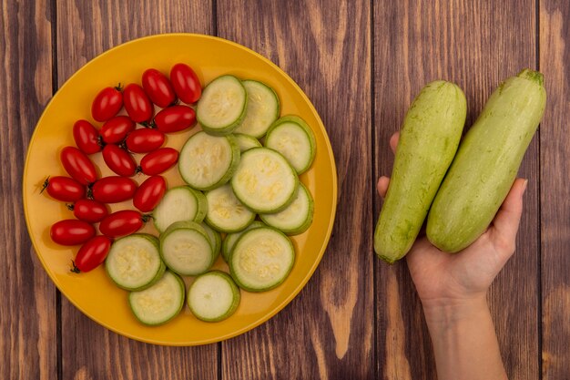 Vista superior de la mano femenina sosteniendo calabacines frescos con placa amarilla de verduras frescas como tomates y calabacines en una pared de madera