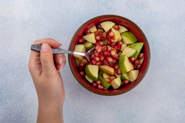 Vista superior de la mano femenina sosteniendo una cuchara con semillas de granada y manzanas picadas en un recipiente rojo sobre blanco