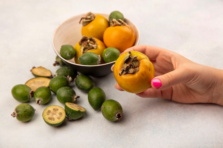 Foto gratuito vista superior de la mano femenina sosteniendo una fruta de caqui maduro naranja con feijoas y caquis aislado sobre una superficie gris
