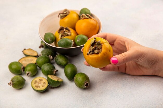 Vista superior de la mano femenina sosteniendo una fruta de caqui maduro naranja con feijoas y caquis aislado sobre una superficie gris