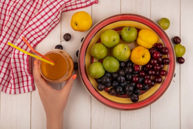 Vista superior de la mano femenina sosteniendo jugo de durazno fresco con un tazón con frutas frescas como cereza verde ciruela cerezas rojas melocotones dulces sobre un fondo blanco de madera