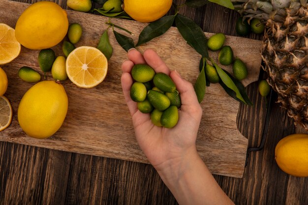 Vista superior de la mano femenina sosteniendo kinkans frescos en una tabla de cocina de madera con limones y kinkans aislado en una pared de madera