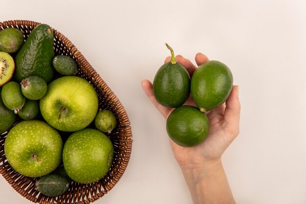 Vista superior de la mano femenina sosteniendo limas frescas con un balde de frutas frescas como manzanas, feijoas y aguacate sobre una superficie blanca