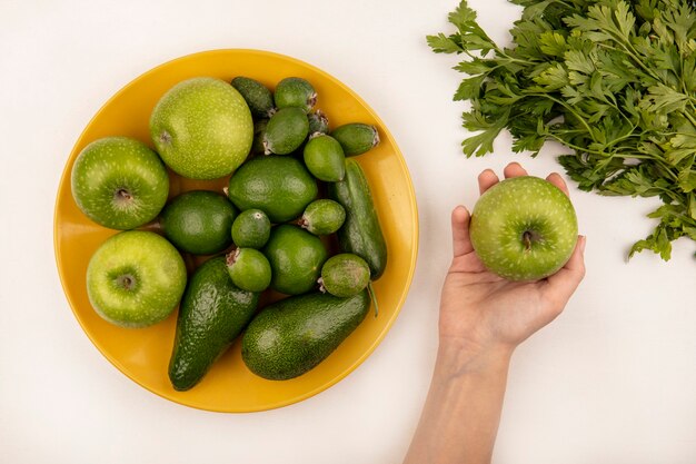 Vista superior de la mano femenina sosteniendo una manzana con plato amarillo de frutas frescas como manzanas, feijoas y aguacate sobre una superficie blanca