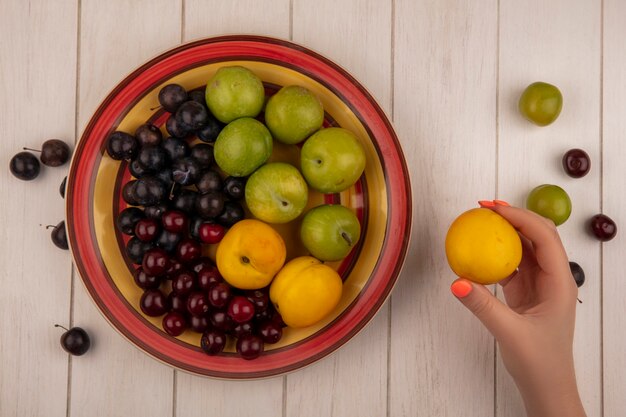 Vista superior de la mano femenina sosteniendo melocotón con un tazón con frutas frescas como cereza verde ciruela cerezas rojas melocotones dulces sobre un fondo blanco de madera