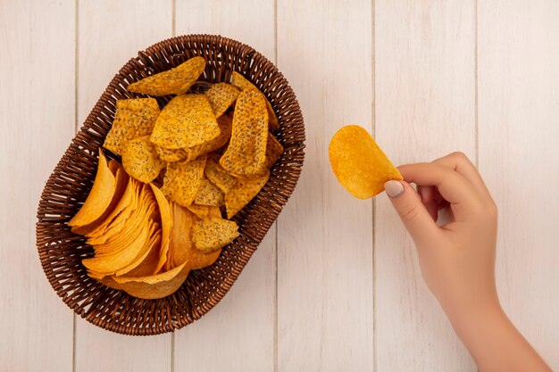 Vista superior de la mano femenina sosteniendo patatas fritas crujientes con un cubo de patatas fritas picantes en una mesa de madera beige