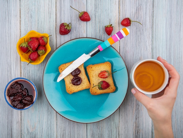 Vista superior de la mano femenina sosteniendo una taza de té con mermelada de fresa con fresas frescas sobre un fondo de madera gris