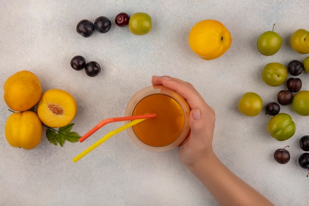 Vista superior de la mano femenina sosteniendo un vaso de jugo de durazno con duraznos frescos con ciruelas cereza verde aislado sobre un fondo blanco.