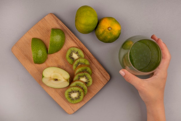 Vista superior de la mano femenina sosteniendo un vaso de jugo de kiwi fresco con rodajas de kiwi de manzanas picadas en una tabla de cocina de madera con mandarinas aisladas