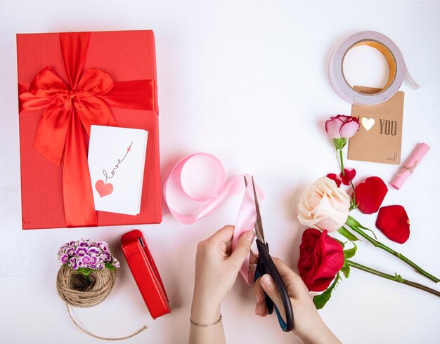 Vista superior de la mano femenina con tijeras corta una cinta rosa y rosas de color rojo y blanco con una caja de regalo roja con un lazo sobre fondo blanco.