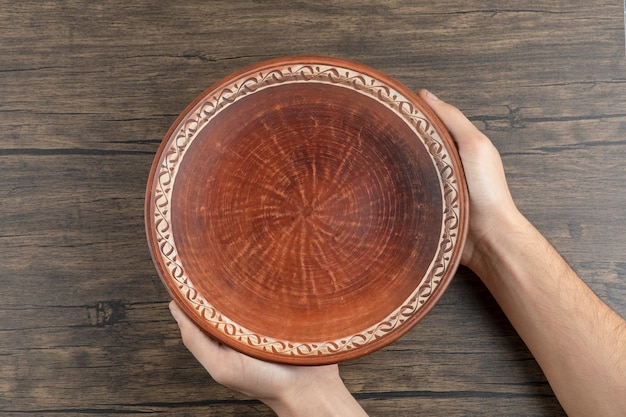 Foto gratuita vista superior de la mano del hombre sosteniendo un plato marrón vacío sobre una mesa de madera.