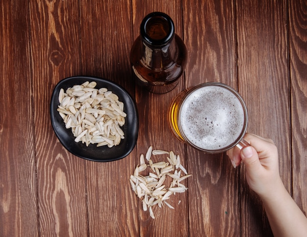 Vista superior de una mano con una jarra de cerveza y una botella de cerveza con aperitivo salado semillas de girasol en un recipiente sobre madera rústica