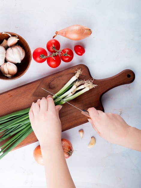 Vista superior de la mano de la mujer cortando la cebolla en la tabla de cortar con tomate ajo sobre fondo blanco.
