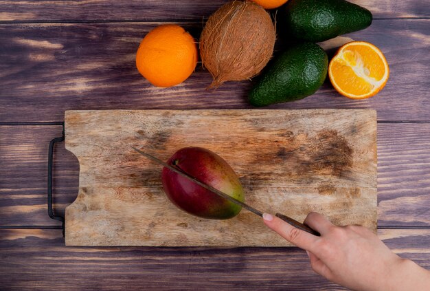 Vista superior de la mano de mujer cortando mango con cuchillo en la tabla de cortar y aguacate de mandarina de coco sobre fondo de madera