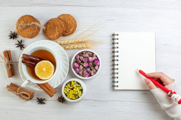 Vista superior de la mano de mujer escribiendo en té negro portátil con limón limón canela y varias galletas herbales sobre fondo blanco.