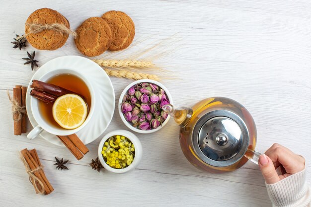 Vista superior de la mano de la mujer sosteniendo una olla con té negro con limón limón canela y varias galletas de hierbas sobre fondo blanco.