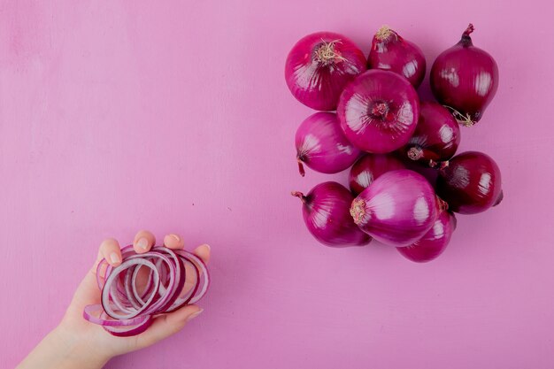 Vista superior de la mano de mujer sosteniendo rodajas de cebolla con cebollas enteras sobre fondo morado con espacio de copia