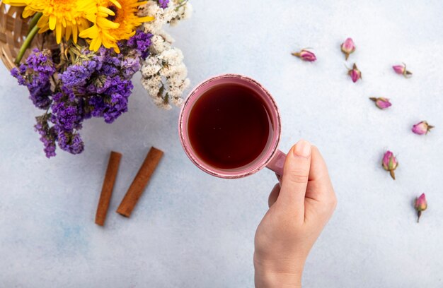 Vista superior de la mano de mujer sosteniendo una taza de té con canela y flores sobre una superficie blanca