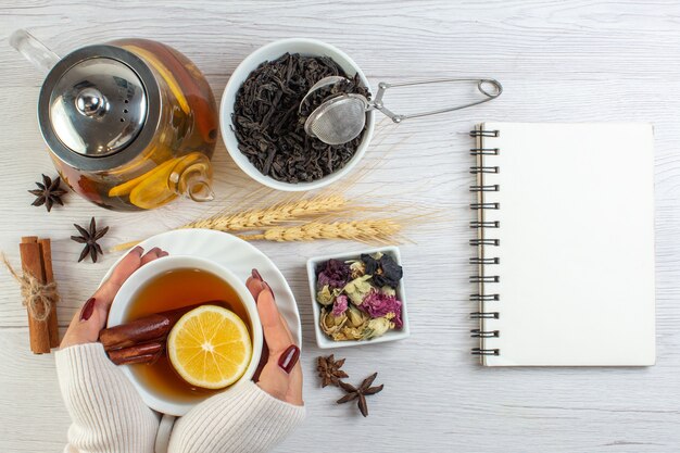 Vista superior de la mano de la mujer sosteniendo una taza con té de hierbas con limón limón canela e ingredientes necesarios junto al cuaderno sobre fondo blanco.