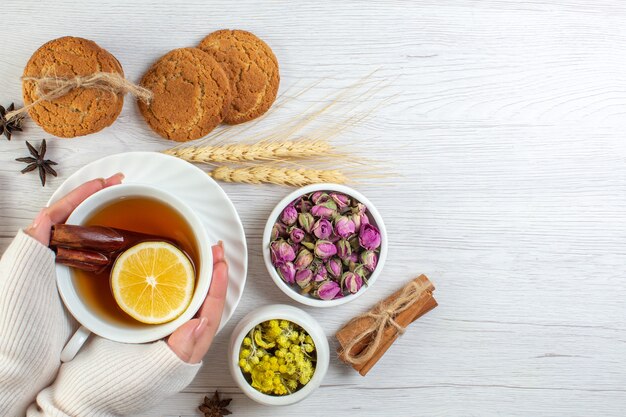 Vista superior de la mano de la mujer sosteniendo una taza con té negro con limón limón canela y varias galletas de hierbas en el lado derecho sobre fondo blanco.