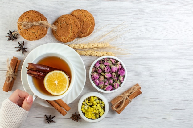 Vista superior de la mano de la mujer sosteniendo una taza con té negro con limón limón canela y varias galletas de hierbas sobre fondo blanco.