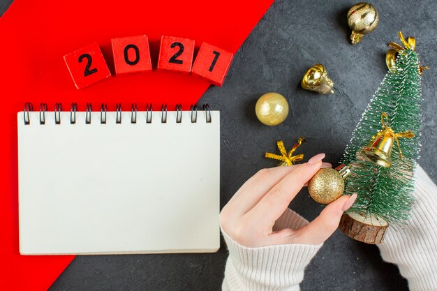 Vista superior de la mano que sostiene el árbol de navidad y accesorios de decoración con números y cuaderno en la mesa oscura