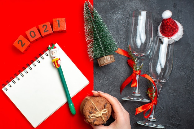 Vista superior de la mano que sostiene el cuaderno espiral y los números de copas de vidrio del árbol de navidad mano que sostiene las galletas apiladas sobre fondo rojo y oscuro