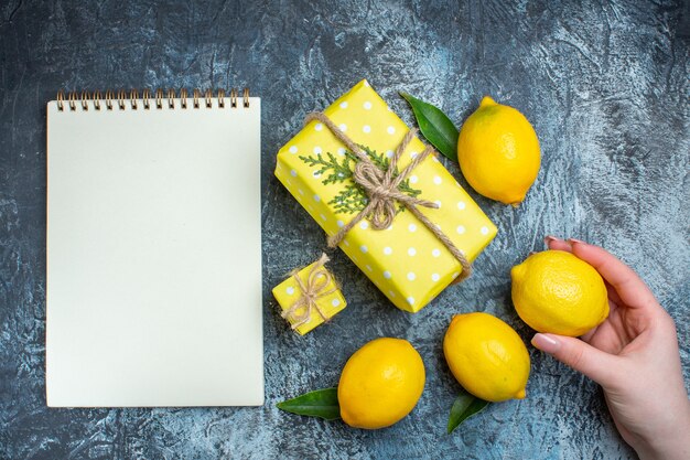 Vista superior de la mano que sostiene uno de los limones frescos con hojas y cajas de regalo amarillas siguiente cuaderno sobre fondo oscuro