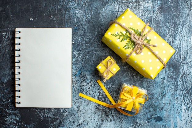 Vista superior de la mano que sostiene una pequeña caja de regalo y otras dos cajas de regalo de Navidad junto al cuaderno de espiral sobre fondo oscuro