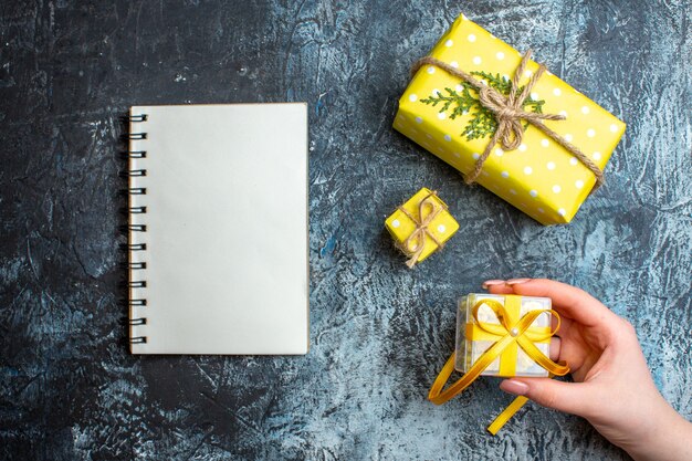 Vista superior de la mano que sostiene una pequeña caja de regalo y otras dos cajas de regalo de Navidad junto al cuaderno sobre fondo oscuro