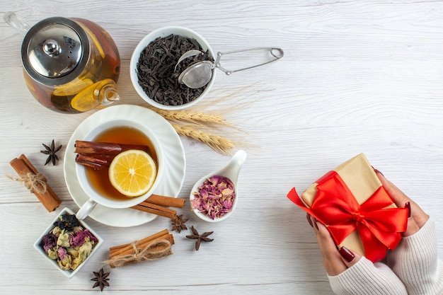 Vista superior de la mano que sostiene los regalos con cinta roja y descanso para el té con canela, lima y limón en una taza y una olla alrededor de los ingredientes necesarios sobre fondo blanco.