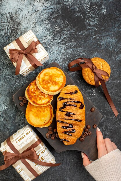 Vista superior de la mano que sostiene la tabla de cortar con un delicioso desayuno con panqueques croisasant galletas apiladas hermosas cajas de regalo en una superficie oscura