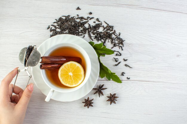 Vista superior de la mano que sostiene el té negro en una taza blanca alrededor de té seco y hojas en la mesa blanca