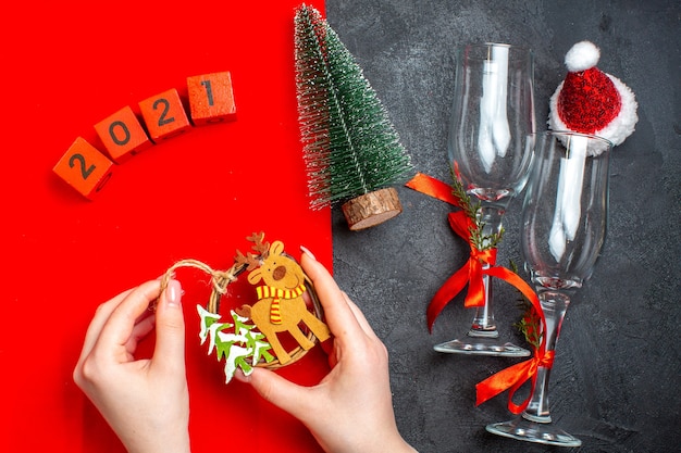 Vista superior de la mano sosteniendo accesorios de decoración copas de vidrio números de árbol de navidad sombrero de santa claus sobre fondo rojo y negro