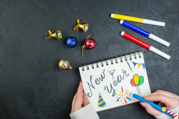 Vista superior de la mano sosteniendo un bolígrafo en un cuaderno de espiral con accesorios de decoración de dibujos y escritura de año nuevo sobre fondo negro