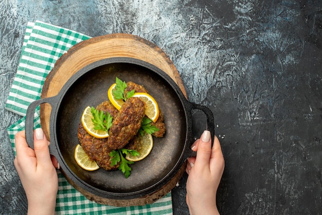 Vista superior de la mano sosteniendo deliciosas chuletas servidas con verduras y limón en una sartén negra sobre una tabla de madera sobre una toalla verde pelada sobre fondo de color oscuro