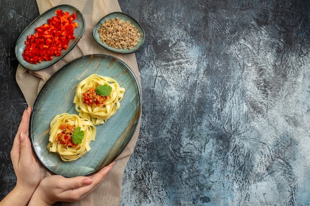 Vista superior de la mano sosteniendo un plato azul con una deliciosa comida de pasta servida con tomate y carne para la cena en una toalla de color bronceado con sus ingredientes