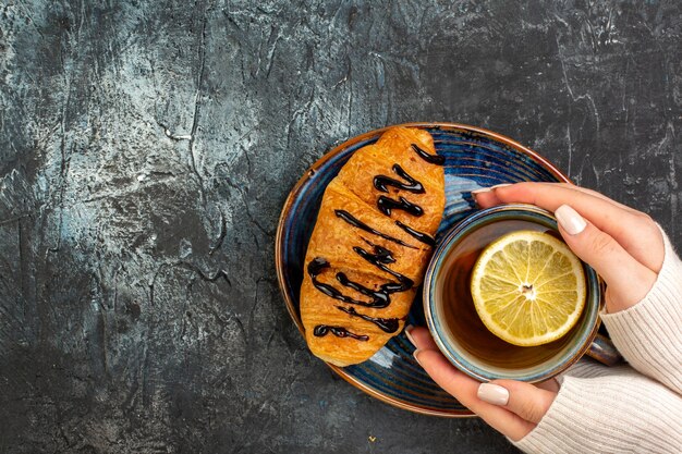 Vista superior de la mano sosteniendo una taza de té negro delicioso croisasant en el lado izquierdo de la mesa oscura