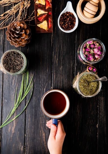 Vista superior de una mano sosteniendo una taza de té y varias especias y hierbas en madera negra con espacio de copia