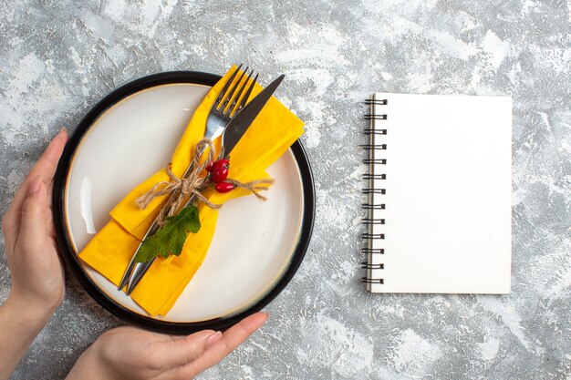 Vista superior de la mano sujetando cubiertos para comida en un plato blanco y cuaderno cerrado sobre la superficie del hielo