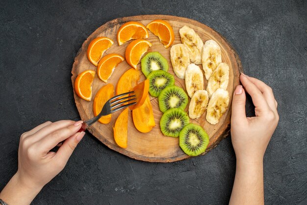 Vista superior de la mano tomando frutas frescas orgánicas naturales con un tenedor en la tabla de cortar sobre fondo oscuro