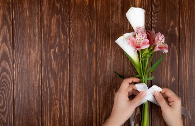 Vista superior de las manos atando con una cinta un ramo de flores de alstroemeria y lirios de color rosa y blanco sobre fondo de madera con espacio de copia
